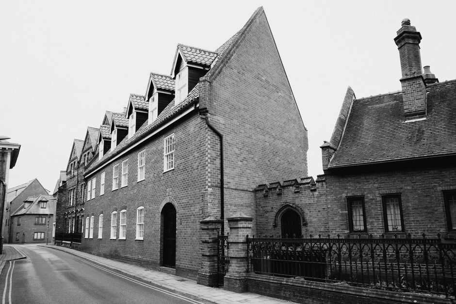 A black and white image depicting a narrow street with an old brick Victorian townhouse in Guildford, featuring a steeply pitched roof with multiple dormer windows and a tall chimney. The building has