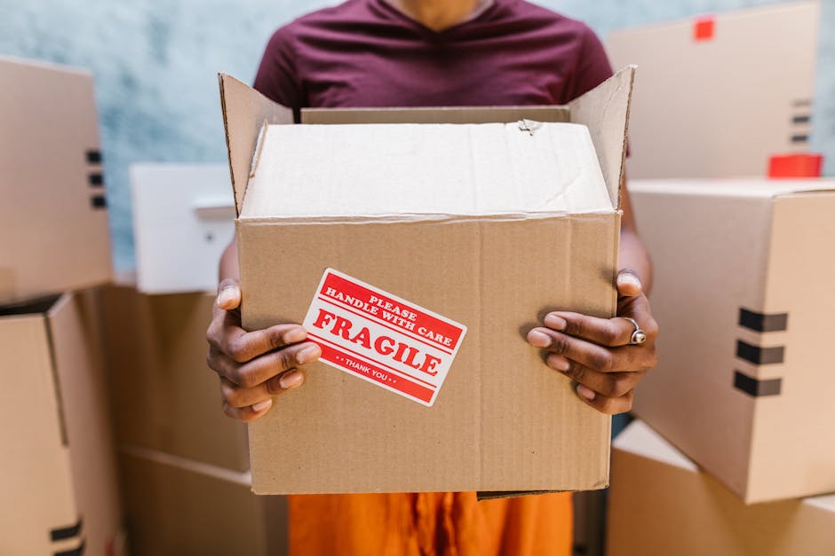 Close-up of a person wearing a dark green shirt and blue pants, using both hands to carefully hold and position a large cardboard box during a home relocation. The box has a white and red caution labe