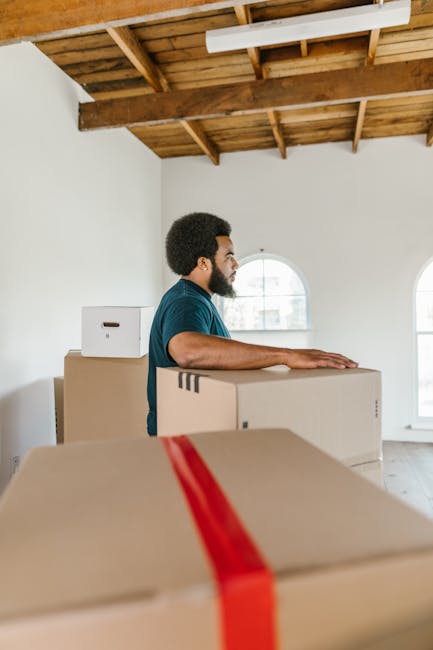 Inside a residential property during a house move, a woman with shoulder-length dark hair and wearing a white top is sitting on the floor, handling a cardboard box that appears to contain packing mate