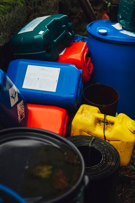 A person wearing a white protective suit, safety goggles, and a black respirator mask stands outdoors in front of a large pile of mixed waste, including plastic bags, cardboard, and debris, on a landf