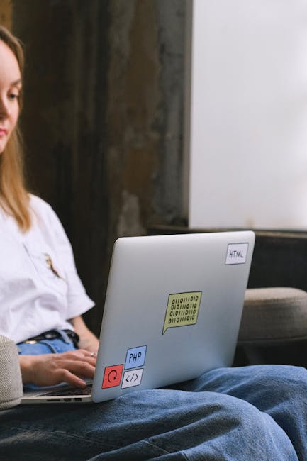 The image shows a young woman sitting on a dark-colored sofa in an indoor setting, with her left arm resting on her lap and her right hand placed on the laptop keyboard. The laptop is open on her lap,