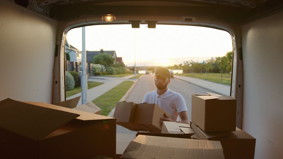 Two men engaged in a home relocation process are seen lifting a large cardboard box from the back of a white moving van parked on a paved driveway, with the van's rear doors open revealing several mor